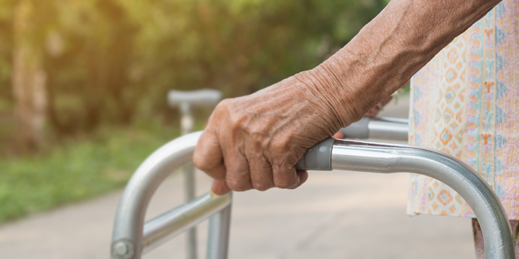 An elderly woman is holding onto her walking frame. Only her hands are in the frame, the background is a leafy green pathway. She is wearing a patterned shirt.