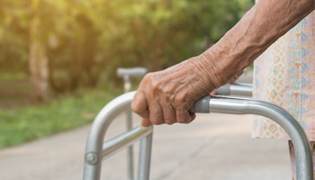 Senior woman holds on to a walking frame while standing on a leafy pathway outdoors.