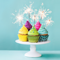 Image of five cupcakes with sparkler candles in them. They are brightly coloured and sitting on a cake stand in front of a blue background.
