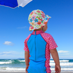 a toddler looks at the ocean dressed in a sun hat, rash shirt, and standing under an umbrella
